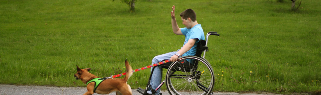 Bilde av en ung gutt i rullestol som er på treningstur med en hund i bånd. Hunden løpet ved siden av gutten.