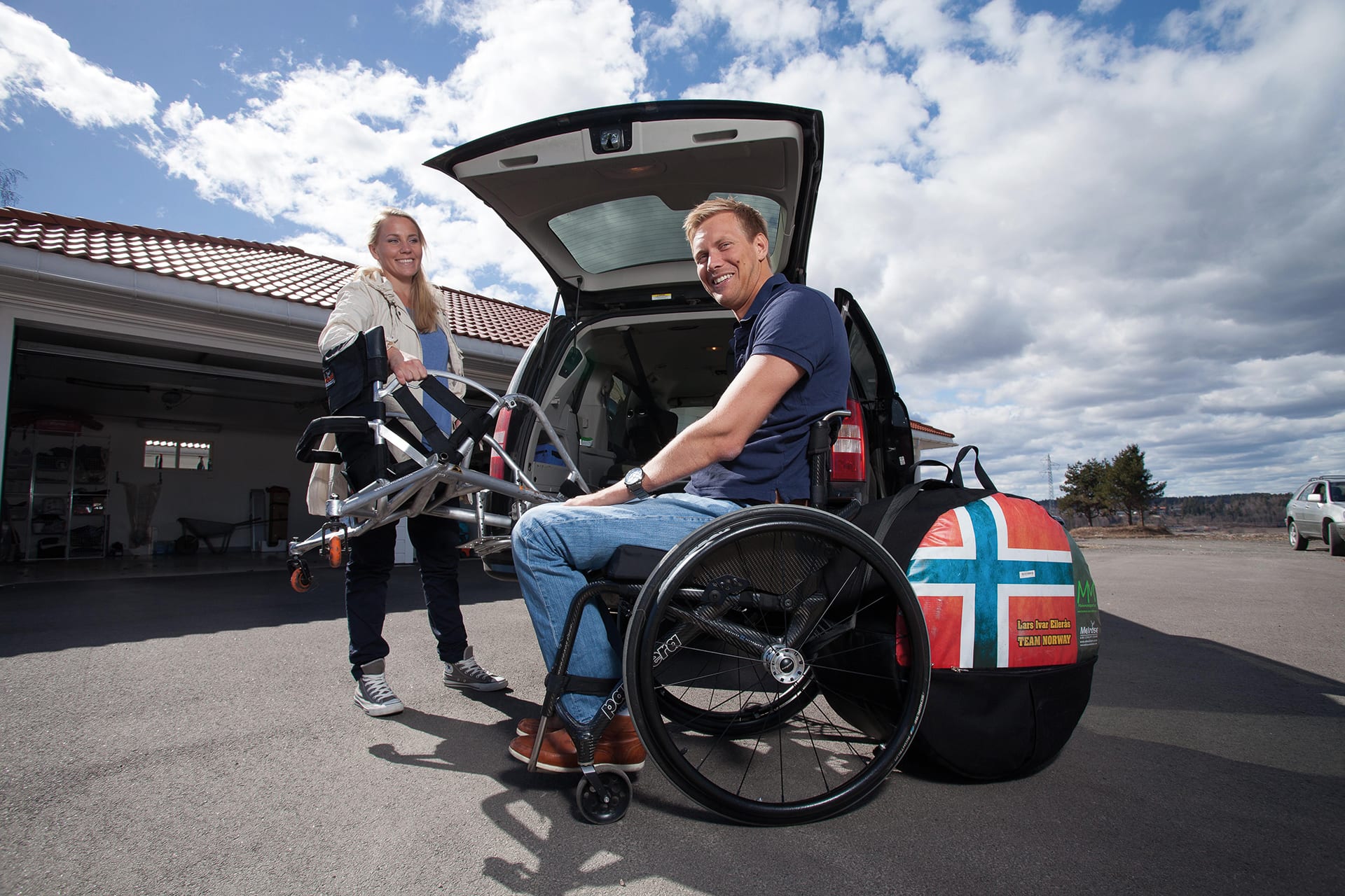 Bilde av artikkelskribent og hans kvinnelige assistent. Han sitter i rullestol foran et åpent bagasjerom til en personbil og smiler til kamera. Bak rullestolen står det som ser ut til å være bagasje. Ved siden av bilen står assistenten. Hun smiler til kamera mens hun hjelper med å plassere utstyr i bilen.