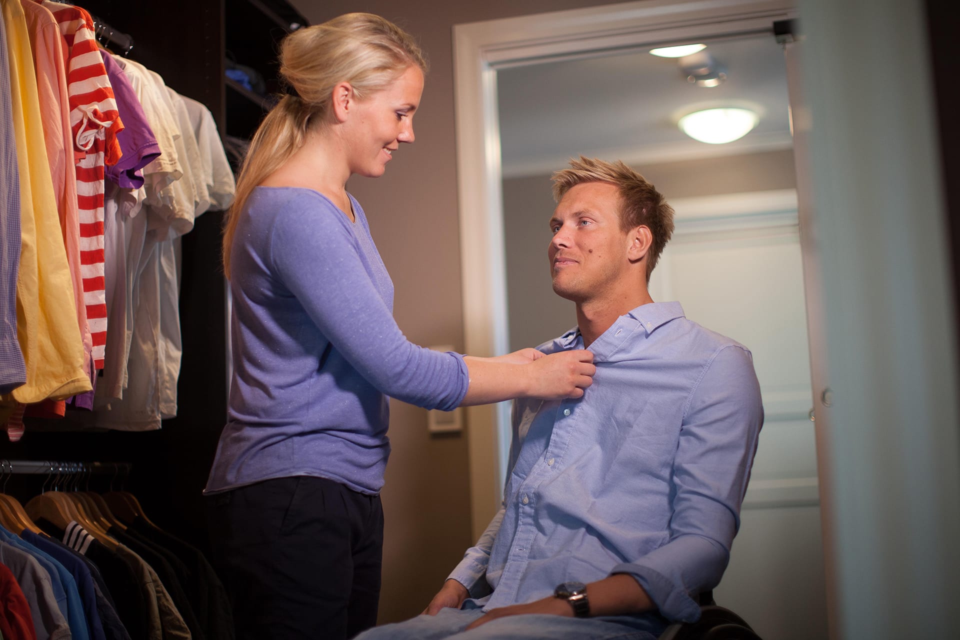 Bilde av artikkelskribent og hans kvinnelige assistent. Han sitter i rullestol mens han kikker mot assistenten. Assistenten hjelper ham med å kneppe igjen en blå skjorte. De befinner seg i et rom med en rekke t-skjorter og skjorter på venstre side.